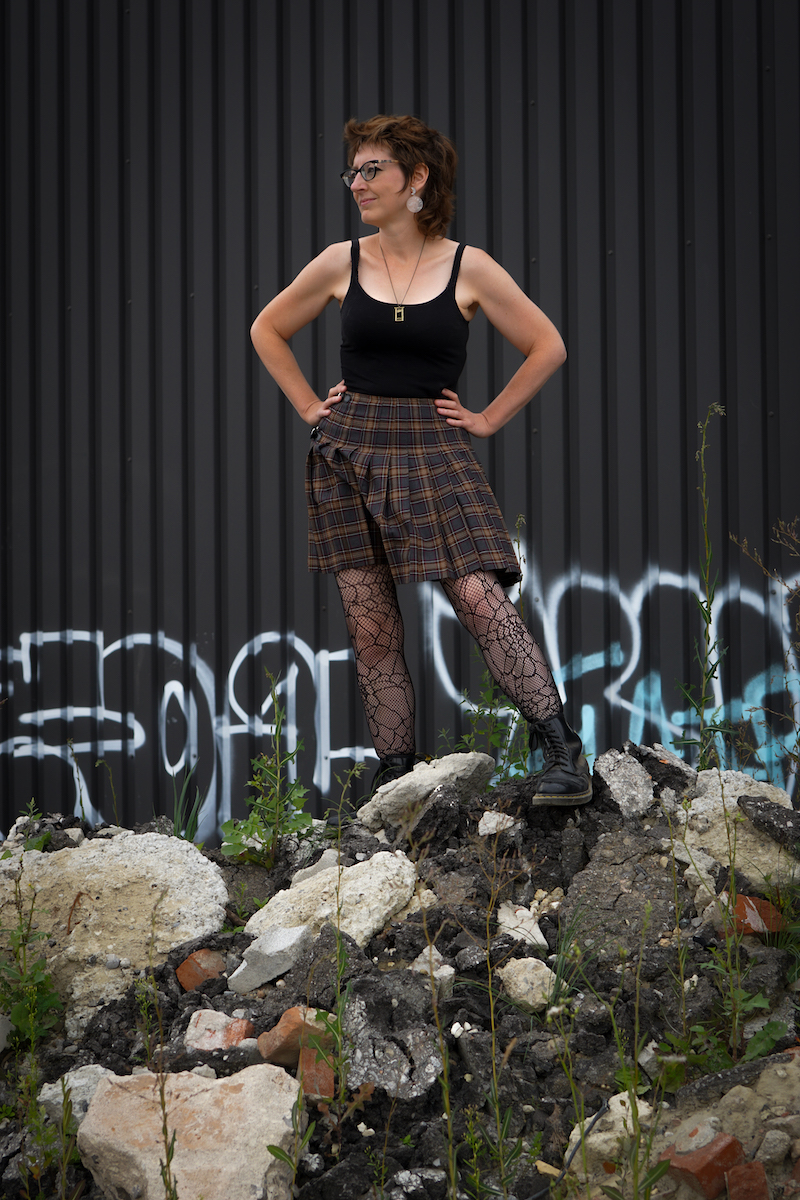 it's a me! Middle aged white woman with a punk haircut in a tanktop and kilt, standing on a pile of rubble.